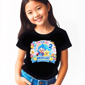May include: A black short-sleeved t-shirt featuring a colorful graphic of the character Plim Plim, a rainbow, balloons, and a train. The text reads PLIM PLIM SAY... 2, BRIELLIE!, and YOU ARE SO SPECIAL! in a bright, playful, multi-colored design.