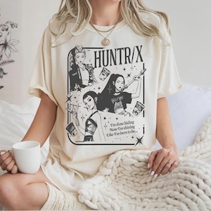 May include: A cream-coloured t-shirt with a black and white graphic design. The design features the text "HUNTR/X" and illustrations of women. The design includes the text "I'm done hiding, Now I'm shining, Like I'm born to be..."