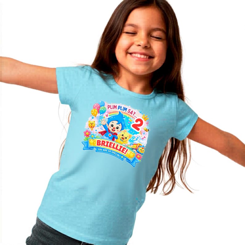 May include: A light blue short-sleeved t-shirt featuring a colorful graphic print of the character Plim Plim, a rainbow, balloons, and the text "PLIM PLIM SAY... 2", "BRIELLIE!", and "YOU ARE SO SPECIAL!" centered on the front of the garment.