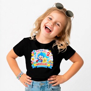 May include: A black short-sleeved t-shirt featuring a colorful graphic print of the cartoon character Plim Plim, balloons, flowers, and the text "PLIM PLIM SAY... 2", "BRIELLIE!", and "YOU ARE SO SPECIAL!" in a bright, festive design.