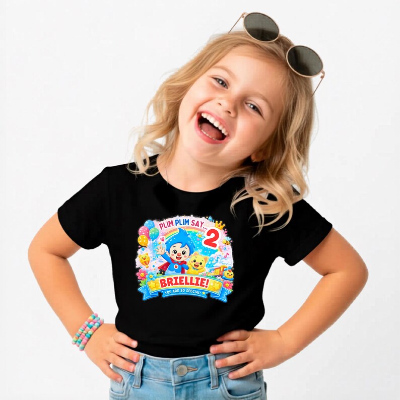 May include: A black short-sleeved t-shirt featuring a colorful graphic print of the cartoon character Plim Plim, balloons, flowers, and the text "PLIM PLIM SAY... 2", "BRIELLIE!", and "YOU ARE SO SPECIAL!" in a bright, festive design.