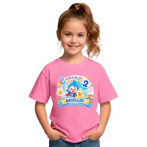 May include: A pink short-sleeved t-shirt featuring a colorful graphic of the cartoon character Plim Plim, a cat, balloons, and a rainbow. The text reads PLIM PLIM SAY... 2 BRIELLIE! YOU ARE SO SPECIAL! The design includes stars and musical notes.