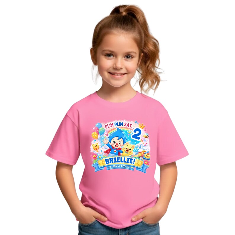 May include: A pink short-sleeved t-shirt featuring a colorful graphic of the cartoon character Plim Plim, a cat, balloons, and a rainbow. The text reads PLIM PLIM SAY... 2 BRIELLIE! YOU ARE SO SPECIAL! The design includes stars and musical notes.