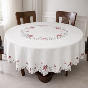 Op de afbeelding: Een ronde witte tafelkleed met een bloemenontwerp. Het tafelkleed heeft een geschulpte rand en is voorzien van geborduurde roze bloemen en zwarte details. De tafel is gedekt met twee houten stoelen.