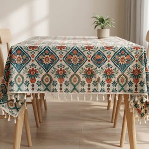Könnte beinhalten: Rechteckige Tischdecke mit cremefarbenem Hintergrund und einem sich wiederholenden Muster aus teal, orangefarbenen und roten Blumen- und geometrischen Designs. Die Tischdecke hat eine Fransenborte und ist über einem Holztisch drapiert. Der Tisch ist von Holzstühlen umgeben.