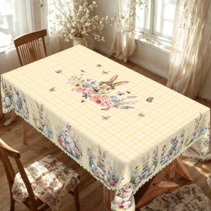 May include: A rectangular yellow gingham tablecloth with a floral and bunny design. The centre features a bunny wearing a floral crown. The border has bunnies, flowers, and butterflies. The tablecloth is on a wooden table.