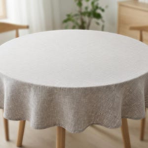 Op de afbeelding: Een ronde, lichtbeige tafelkleed met een getextureerde weefsel, gedrapeerd over een houten tafel. De tafel heeft vier slanke, lichtgekleurde houten poten. Het tafelkleed is gemaakt van een linnenachtig materiaal.