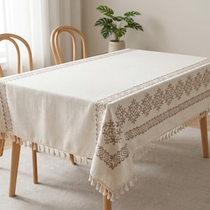 Op de afbeelding: Een rechthoekig tafelkleed in gebroken wit met een bruine geometrische rand en kwastjes. Het tafelkleed ligt op een houten tafel. De tafel is ongeveer 150 cm lang. Twee houten stoelen zijn zichtbaar.