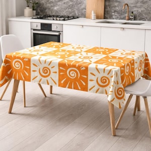 May include: A rectangular orange and white tablecloth with a repeating sun design. The tablecloth is on a wooden-legged table, with two white chairs. The kitchen has white cabinets and a stainless steel oven.