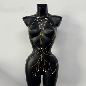 May include: Gold-toned body chain with delicate strands and small, white pearl accents. The jewelry piece is displayed on a black mannequin, showcasing its design and draping. The body chain is a fashion accessory.