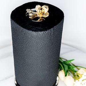 May include: A black paper towel holder with a gold floral accent on top and bottom. The paper towel roll is black and has a textured pattern. The holder has a silver base and a gold floral design.