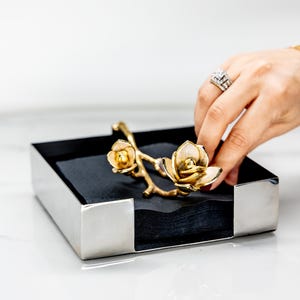 May include: A silver napkin holder with a black interior, featuring a gold floral accent. The decorative piece has a rectangular shape with a polished finish. The floral accent is a gold-colored branch with two flower blossoms.