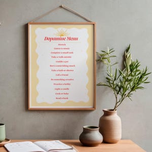 Peut inclure: Une affiche encadrée intitulée "Dopamine Menu" avec une liste d'activités. L'affiche a un fond jaune clair et un cadre en bois, accrochée à un mur gris. Un vase avec de la verdure et deux vases en céramique sont au premier plan.