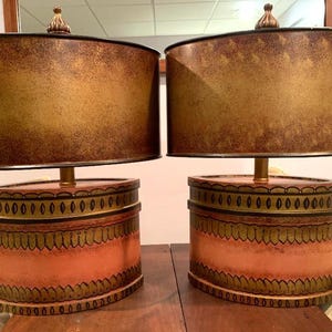 May include: Two matching table lamps with oval shades and decorative bases. The shades are a mottled gold and brown, and the bases are peach with gold and black trim. Each lamp has a gold-colored finial.