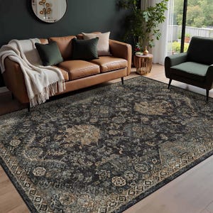 Könnte beinhalten: Ein großer, dunkel gefärbter Teppich mit einem verzierten Blumenmuster. Der Teppich hat einen Used-Look mit Schwarz-, Grau- und Beigetönen. Er befindet sich in einem Wohnzimmer mit einem braunen Ledersofa, einem grünen Sessel und dekorativen Kissen.
