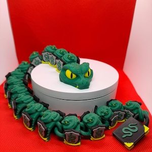 May include: A long, articulated green snake toy with yellow eyes, composed of multiple segments connected by small black shields featuring a green snake emblem. A separate black rectangular book-like object with a green snake symbol is included.
