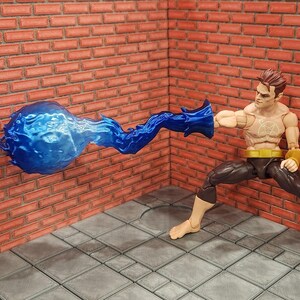May include: A superhero action figure in a dynamic pose, with a blue energy blast emanating from his hand. The figure is wearing black pants, a yellow belt, and wristbands. The background features a brick wall and a tiled floor.