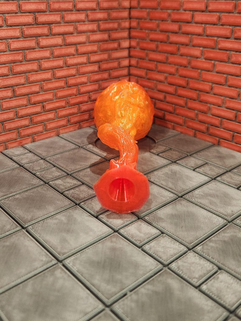May include: An orange, abstract sculpture with a bulbous top and a flared base. The sculpture is made of a translucent material, and the background features a brick wall and a tiled floor.