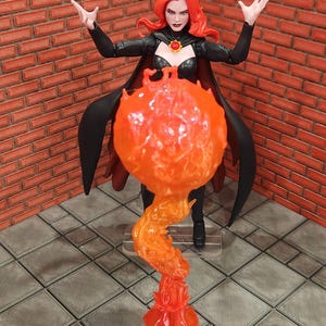 May include: A red-haired action figure in a black outfit with a cape, raising her arms. A translucent orange flame effect emanates from the figure's hands. The figure stands on a gray tiled surface, with a brick wall in the background.