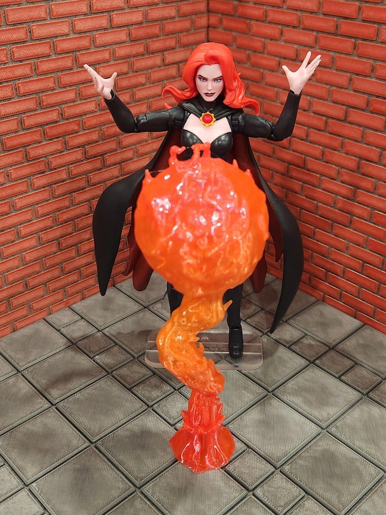 May include: A red-haired action figure in a black outfit with a cape, raising her arms. A translucent orange flame effect emanates from the figure's hands. The figure stands on a gray tiled surface, with a brick wall in the background.