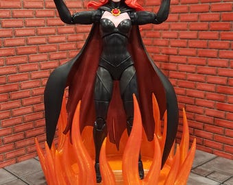 May include: A posable action figure of a character with long red hair, wearing a black bodysuit and a black cape with a red and gold circular brooch. The figure stands inside a translucent orange plastic flame effect base with arms raised.