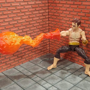 May include: Action figure of a muscular character with brown hair, wearing dark pants and a gold belt. The figure is posed with a fiery orange energy blast emanating from its fist, striking a red brick wall. A symbol is on the figure's chest.