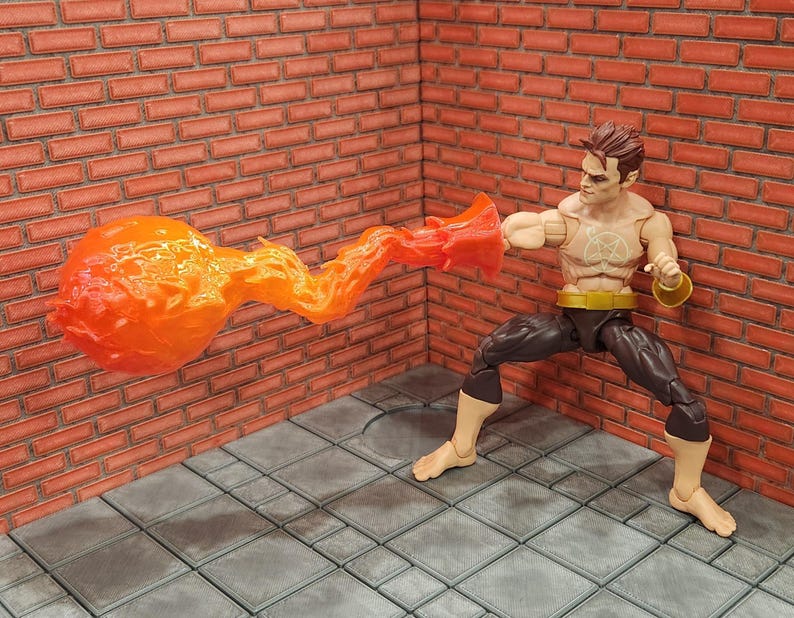 May include: Action figure of a muscular character with brown hair, wearing dark pants and a gold belt. The figure is posed with a fiery orange energy blast emanating from its fist, striking a red brick wall. A symbol is on the figure's chest.