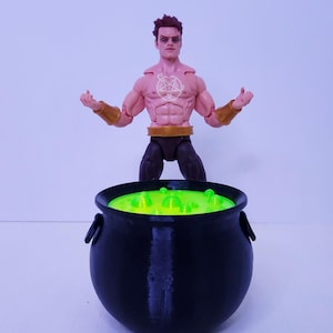 May include: A muscular action figure with brown hair, dark eye makeup, and a white pentagram symbol on the chest, wearing gold wrist cuffs and a belt. The figure stands behind a black cauldron filled with a bright, neon green bubbling liquid.