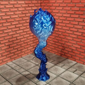 May include: A blue, flame-shaped sculpture with a spherical top and a swirling, stylized base. The sculpture has a glossy finish and is set against a brick wall and tiled floor.