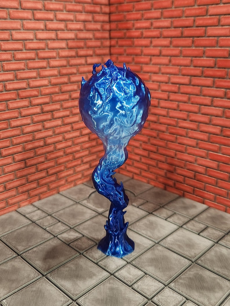 May include: A blue, flame-shaped sculpture with a spherical top and a swirling, stylized base. The sculpture has a glossy finish and is set against a brick wall and tiled floor.