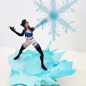 May include: A collectible action figure featuring a character with short black and white hair, wearing a blue crop top, black jacket, and white shorts. The figure stands on a jagged, light blue ice-textured base, holding a large, translucent blue snowflake.