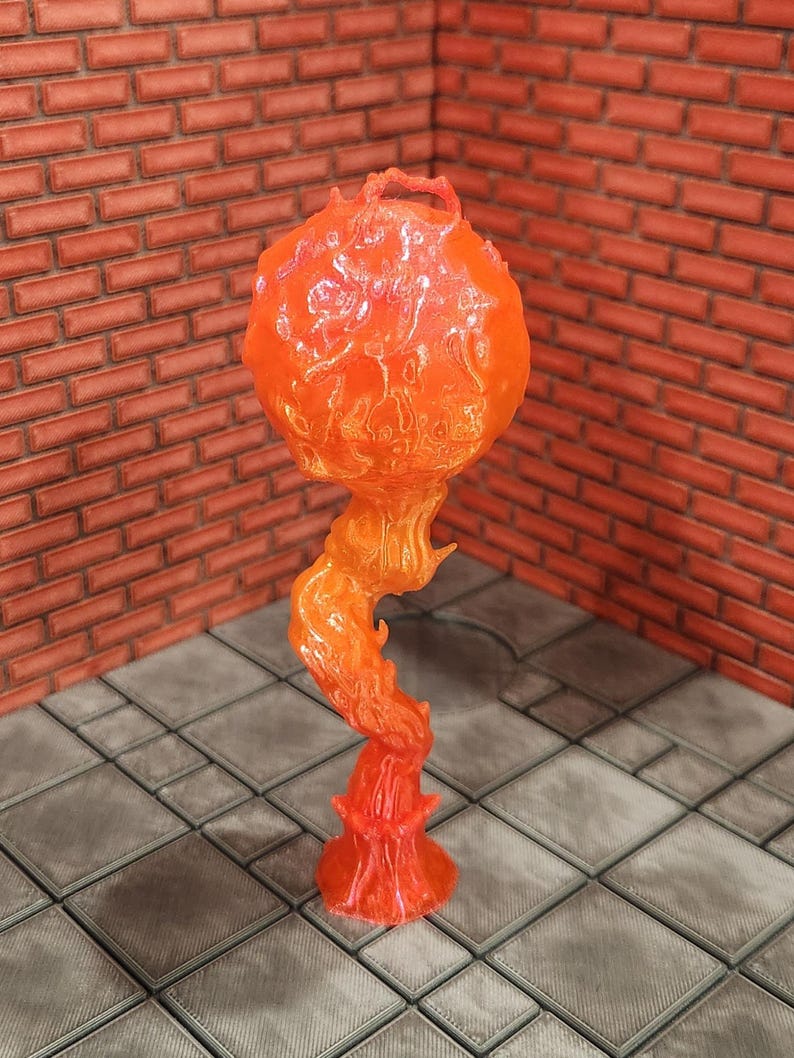 May include: A translucent orange and red decorative sculpture resembling a stylized flame. The sculpture has a bulbous top with a textured surface, supported by a swirling, flame-like stem and a flared base. The piece is set against a brick wall and a tiled floor.