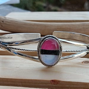 May include: A silver cuff bracelet with an oval-shaped, color-blocked stone. The stone features pink, black, and white sections. The bracelet has a twisted rope design around the stone and along the sides. The text "Silver Artisan2026" is visible.