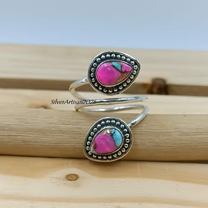 May include: A silver-toned ring with two teardrop-shaped settings. Each setting features a vibrant pink stone with turquoise and gold accents, surrounded by a beaded border. The ring wraps around the finger.