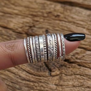 May include: A collection of silver rings with various designs, including twisted rope, floral, and geometric patterns. The rings are stacked on a finger, showcasing the different textures and styles. The rings are made of silver.