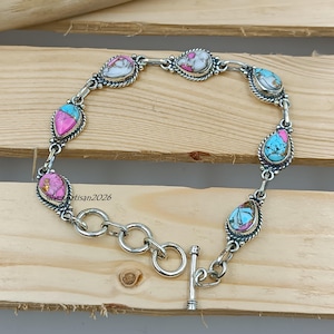 May include: A silver bracelet with teardrop-shaped stones in shades of pink, turquoise, and white. The stones are set in a silver frame with a twisted rope design. The bracelet has a toggle clasp. The text "silver artisan2026" is visible.
