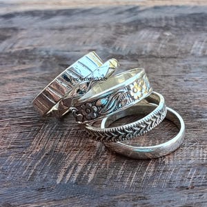 May include: A set of four silver rings stacked together. The rings feature varied designs including a smooth band, a band with a repeating chevron pattern, a band with embossed floral motifs, and a wide band with a textured, hammered finish.