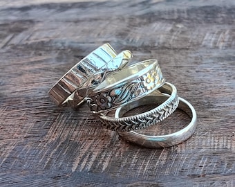 Könnte beinhalten: Ein Set aus vier gestapelten Silberringen. Die Ringe weisen unterschiedliche Designs auf, darunter ein glattes Band, ein Band mit wiederkehrendem Fischgrätenmuster, ein Band mit geprägten Blumenmotiven und ein breites Band mit gehämmertem Finish.