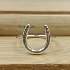 May include: A silver horseshoe ring. The ring features a polished horseshoe design, a symbol of good luck. The ring is set against a wooden background. The text "SilverArtisan2026" is visible on the left side of the image.