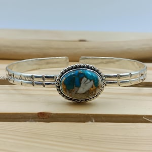 May include: Silver cuff bracelet with an oval turquoise and white stone centerpiece. The stone is framed by a twisted rope design. The silver band has a decorative etched pattern. The bracelet is displayed on a wooden surface.