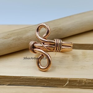 K&ouml;nnte beinhalten: Ein kupferfarbener Ring mit einem einzigartigen, handgefertigten Design. Der Ring hat eine gewickelte Ober- und Unterseite, die durch ein gewickeltes Band verbunden sind. Der Ring wird auf einer Holzoberfl&auml;che pr&auml;sentiert.