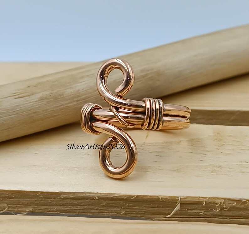 K&ouml;nnte beinhalten: Ein kupferfarbener Ring mit einem einzigartigen, handgefertigten Design. Der Ring hat eine gewickelte Ober- und Unterseite, die durch ein gewickeltes Band verbunden sind. Der Ring wird auf einer Holzoberfl&auml;che pr&auml;sentiert.
