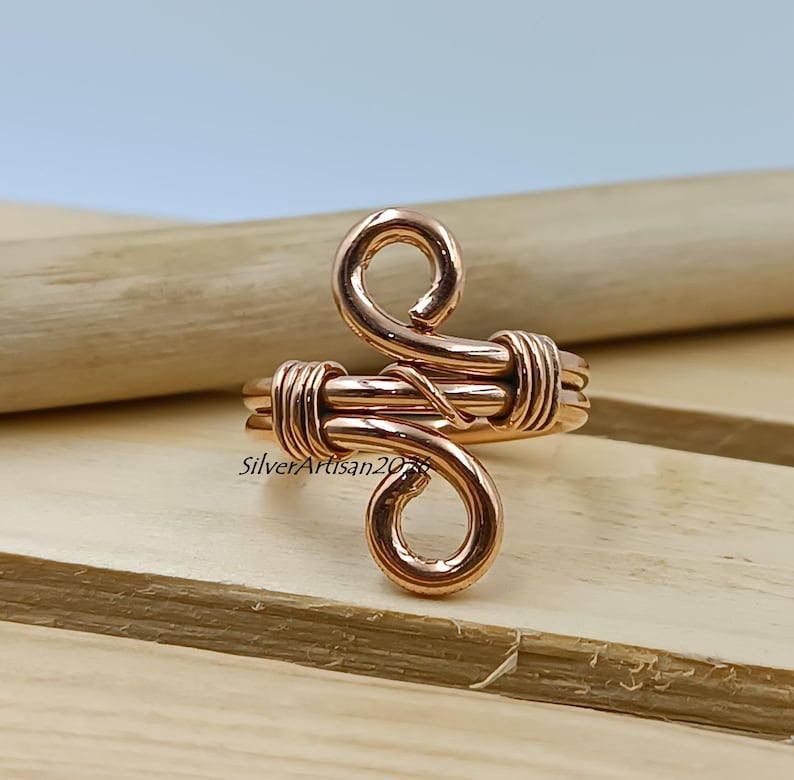K&ouml;nnte beinhalten: Ein kupferfarbener Drahtring mit einem einzigartigen, wirbelnden Design. Der Ring hat zwei gebogene Schlaufen und ein gewickeltes Band, das eine handwerkliche &Auml;sthetik zeigt. Der Ring wird auf einer Holzoberfl&auml;che pr&auml;sentiert.