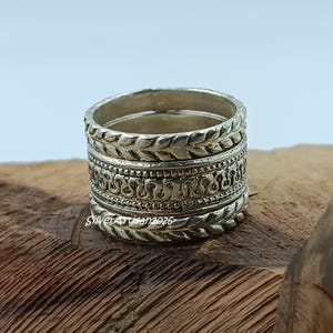Puede incluir: Pila de anillos de plata con diseños intrincados. Los anillos presentan patrones detallados, incluyendo hojas y adornos. Los anillos están apilados sobre una superficie de madera, mostrando la artesanía y la textura de la joyería.