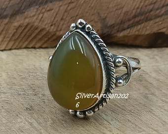May include: A silver ring with a teardrop-shaped, olive-green gemstone. The ring has a detailed, braided silver band and decorative silver accents. The text "SilverArtisan202" and the number "6" are visible on the ring.