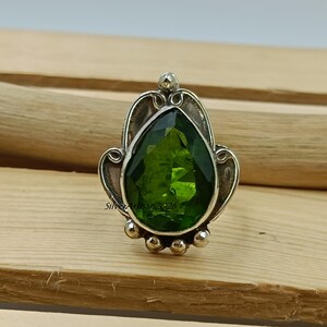 May include: A silver ring featuring a large, pear-shaped, faceted green gemstone. The gemstone is set in a decorative silver setting with ornate details and small silver beads. The ring is displayed on a light brown wooden surface.