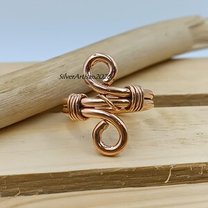 K&ouml;nnte beinhalten: Ein kupferfarbener Drahtring mit einem einzigartigen, wirbelnden Design. Der Ring hat ein gewickeltes Band und zwei dekorative Wirbel. Der Text "SilverArtisan2026" ist auf der Holzoberfl&auml;che sichtbar. Der Ring wird auf einer Holzoberfl&auml;che pr&auml;sentiert.