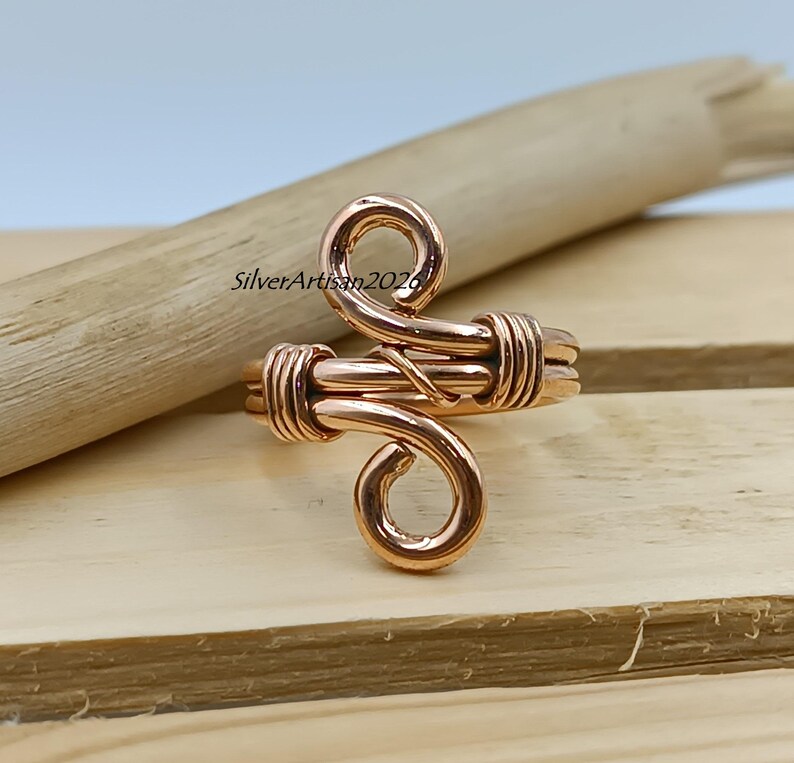 K&ouml;nnte beinhalten: Ein kupferfarbener Drahtring mit einem einzigartigen, wirbelnden Design. Der Ring hat ein gewickeltes Band und zwei dekorative Wirbel. Der Text "SilverArtisan2026" ist auf der Holzoberfl&auml;che sichtbar. Der Ring wird auf einer Holzoberfl&auml;che pr&auml;sentiert.
