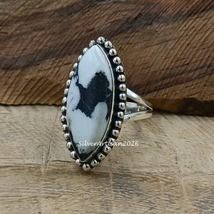 May include: A silver ring with a large, oval white stone with black veining. The stone is framed by a beaded silver border. The ring's band splits into two at the top. The ring is set against a wooden background.