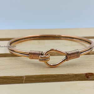 May include: A copper-colored bracelet with a hook clasp. The bracelet is made of two parallel wires, with wire wrapping at the clasp and on each side. The bracelet is resting on a wooden surface.